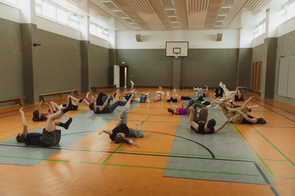 In einer Sporthalle liegen mehrere Personen verteilt auf dem Boden und führen eine Übung mit angehobenen Beinen und Armen aus. Im Hintergrund sind ein Basketballkorb, hohe Fenster und graue Hallenwände zu sehen.