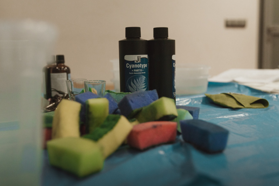 Auf einem mit blauer Folie bedeckten Tisch stehen zwei schwarze Flaschen mit Cyanotypie-Chemikalien neben kleinen Gläsern. Davor liegen bunte Schwämme und Stoffstücke, während der Hintergrund unscharf bleibt.