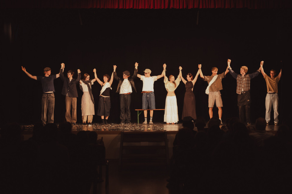Eine Gruppe von Schauspielerinnen und Schauspielern steht auf einer Theaterbühne und verbeugt sich mit erhobenen, ineinandergelegten Händen vor dem Publikum. Der dunkle Bühnenhintergrund, der rote Vorhang und die silhouettenhaften Zuschauer im Vordergrund erzeugen eine feierliche Abschlussstimmung.