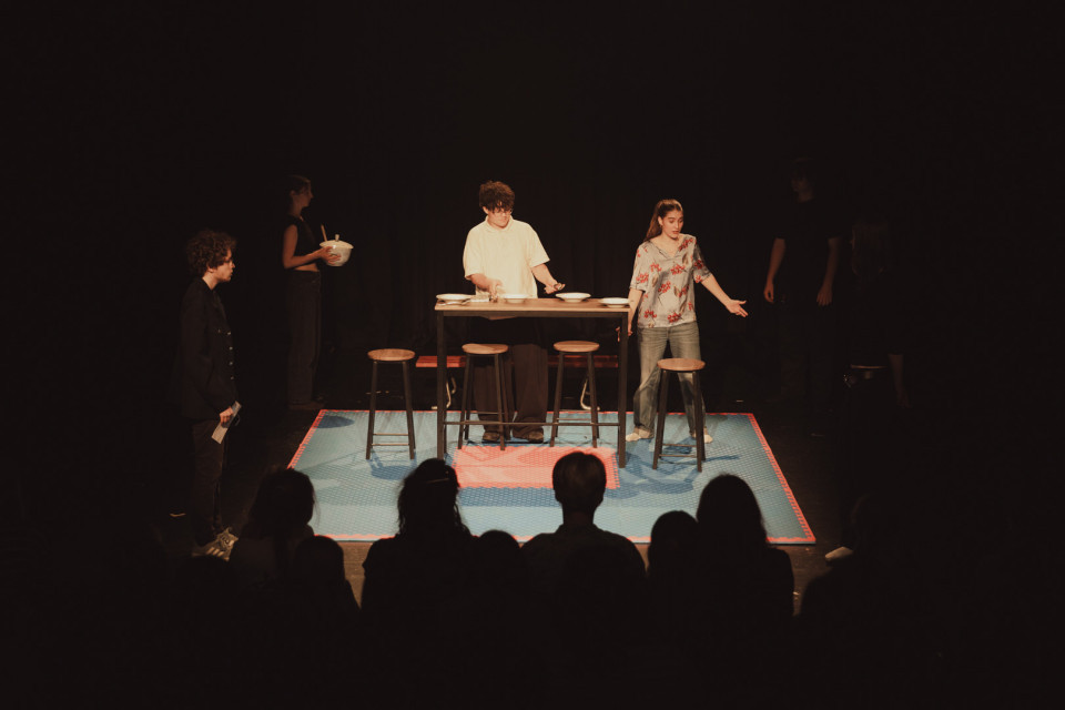 Auf einer dunklen Theaterbühne stehen mehrere Personen um einen hohen Tisch mit Tellern und Hockern, während das Publikum im Vordergrund als Silhouette zu sehen ist. Ein junger Mann in hellem Hemd und eine junge Frau in geblümter Bluse sind im Scheinwerferlicht besonders hervorgehoben.