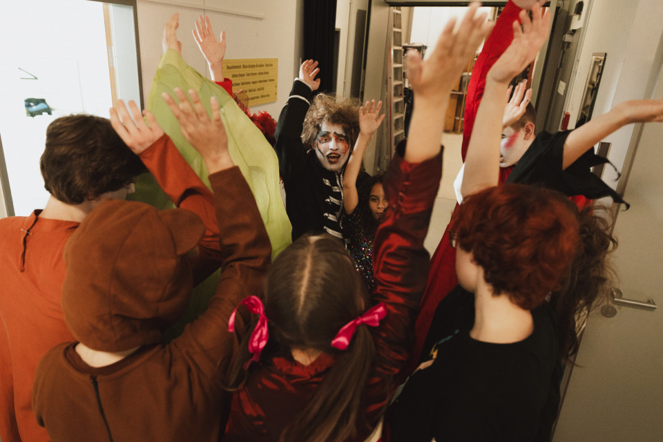 Mehrere Kinder in Kostümen und mit geschminkten Gesichtern stehen in einem Raum eng zusammen und heben die Arme. Die Szene wirkt wie eine fröhliche Theater- oder Faschingsvorbereitung hinter den Kulissen.