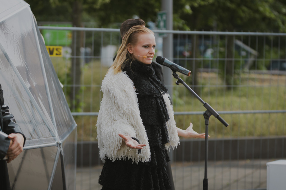 Eine Frau mit blondem Haar steht draußen vor einem Mikrofon und trägt einen auffälligen schwarz-weißen, fransigen Mantel. Im Hintergrund sind Absperrgitter, Bäume und weitere Personen zu sehen, während sie mit ausgebreiteten Händen spricht oder singt.