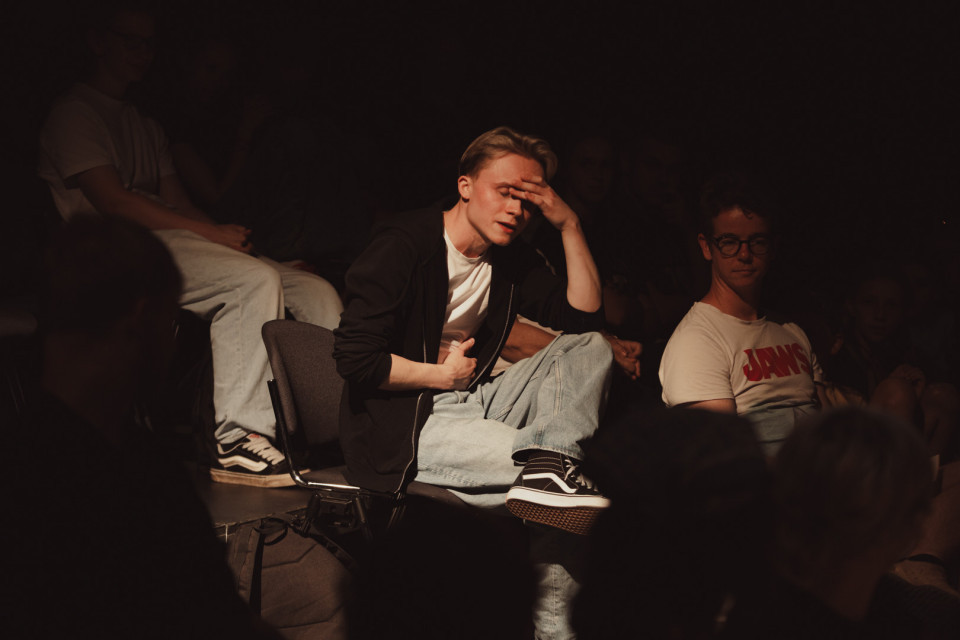 In einem dunklen Raum sitzt ein junger Mann im Scheinwerferlicht auf einem Stuhl und hält sich nachdenklich die Hand an die Stirn. Um ihn herum sitzen weitere Personen im Halbdunkel, wodurch eine konzentrierte, theaterartige Atmosphäre entsteht.