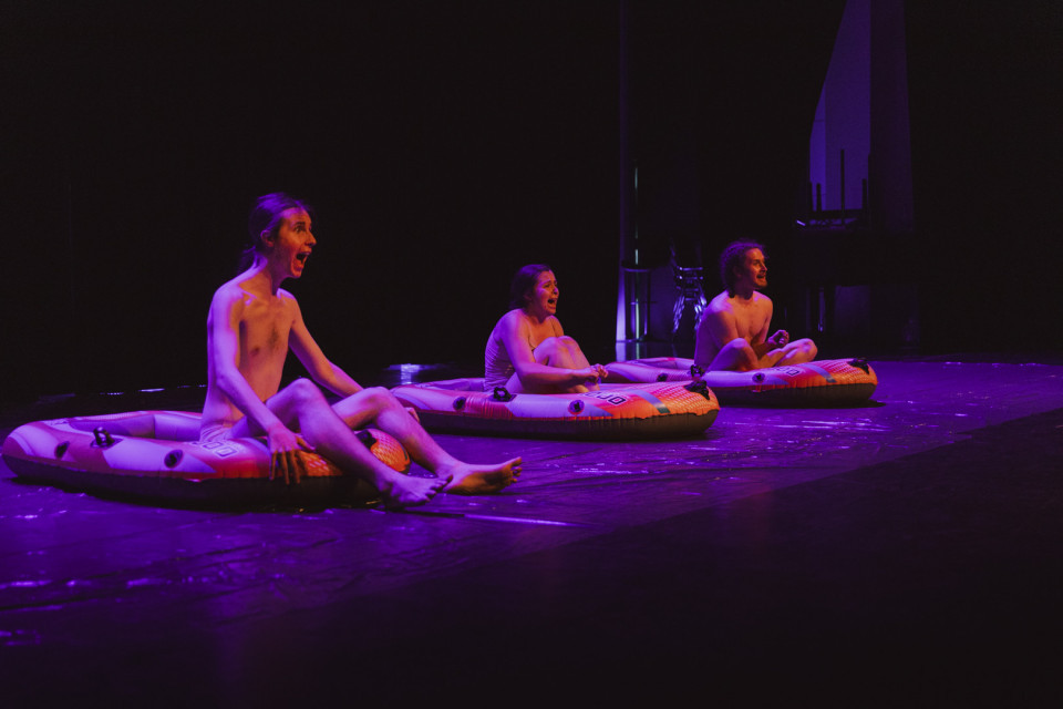 Drei Personen sitzen auf aufblasbaren Schlauchbooten auf einer dunklen Bühne, die in violettes Licht getaucht ist. Sie wirken wie Darsteller in einer Theater- oder Performance-Szene und schauen mit geöffneten Mündern nach vorne.