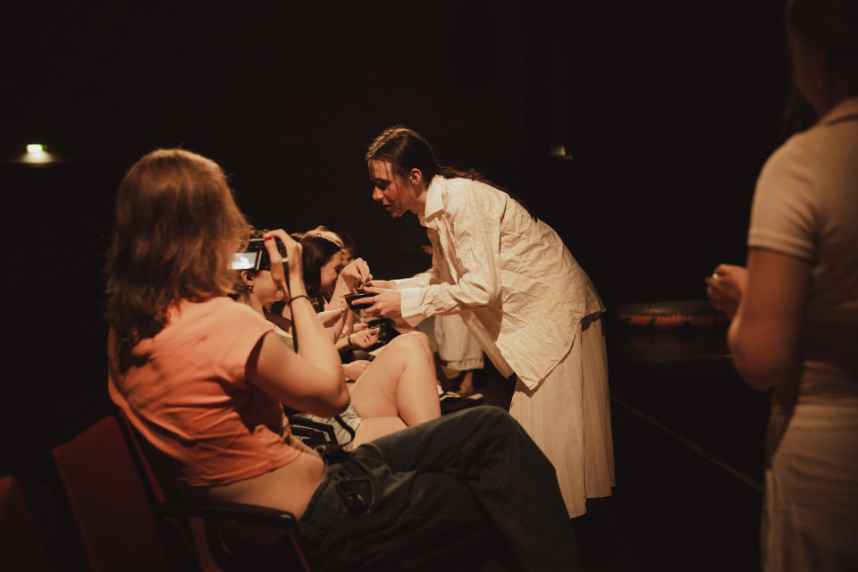 In einem dunkel beleuchteten Theaterraum beugt sich eine Frau in weißer Kleidung zu sitzenden Zuschauerinnen und reicht ihnen etwas. Eine Person im Vordergrund fotografiert die Szene mit einem Smartphone, während rechts unscharf eine weitere Person steht.