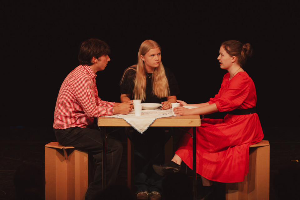 Drei Personen sitzen auf einer Bühne an einem kleinen Tisch und scheinen ein ernstes Gespräch zu führen. Vor ihnen stehen Becher und ein Teller, während der dunkle Hintergrund die Szene wie eine Theateraufführung wirken lässt.