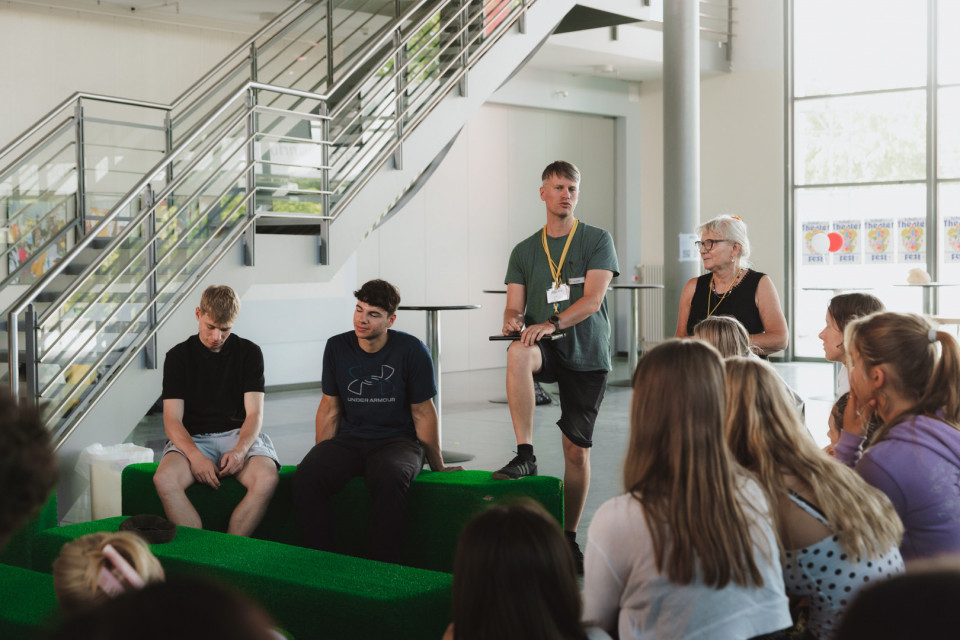 In einem hellen, modernen Foyer mit Treppe sitzt eine Gruppe junger Menschen auf grünen Sitzgelegenheiten und hört zu. Ein Mann mit Namensschild spricht stehend vor der Gruppe, während eine ältere Frau neben ihm steht.