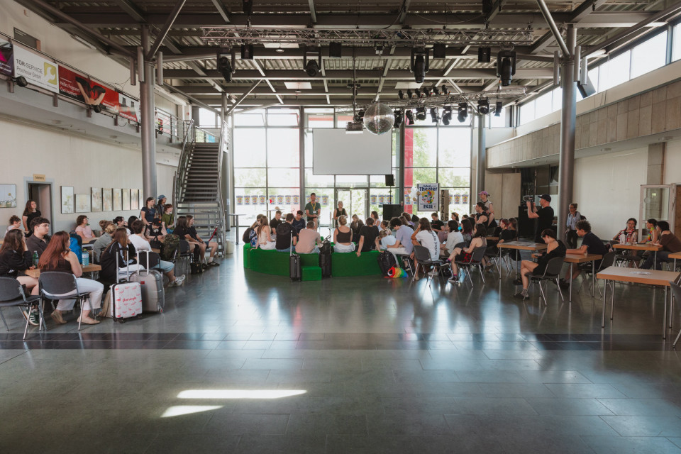In einer großen, hellen Halle sitzen viele Jugendliche und junge Erwachsene in Gruppen auf Stühlen und grünen Sitzblöcken. Über ihnen hängen Bühnenlichter und eine Discokugel, während im Hintergrund große Fenster, eine Treppe und Plakate zu sehen sind.