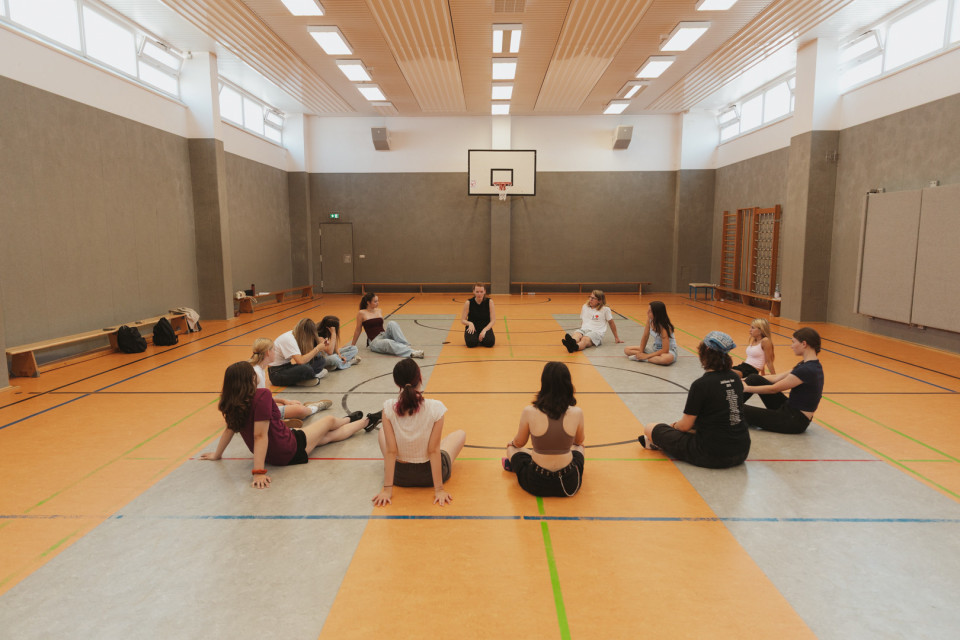 Eine Gruppe Jugendlicher sitzt in einer Sporthalle im Kreis auf dem Boden. Im Hintergrund sind ein Basketballkorb, Bänke und Turngeräte zu sehen.
