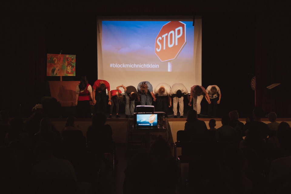 Auf einer Bühne verbeugen sich mehrere Personen vor einem Publikum in einem abgedunkelten Saal. Im Hintergrund ist eine große Projektion mit einem Stoppschild und dem Hashtag „#blockmichnichtklein“ zu sehen.