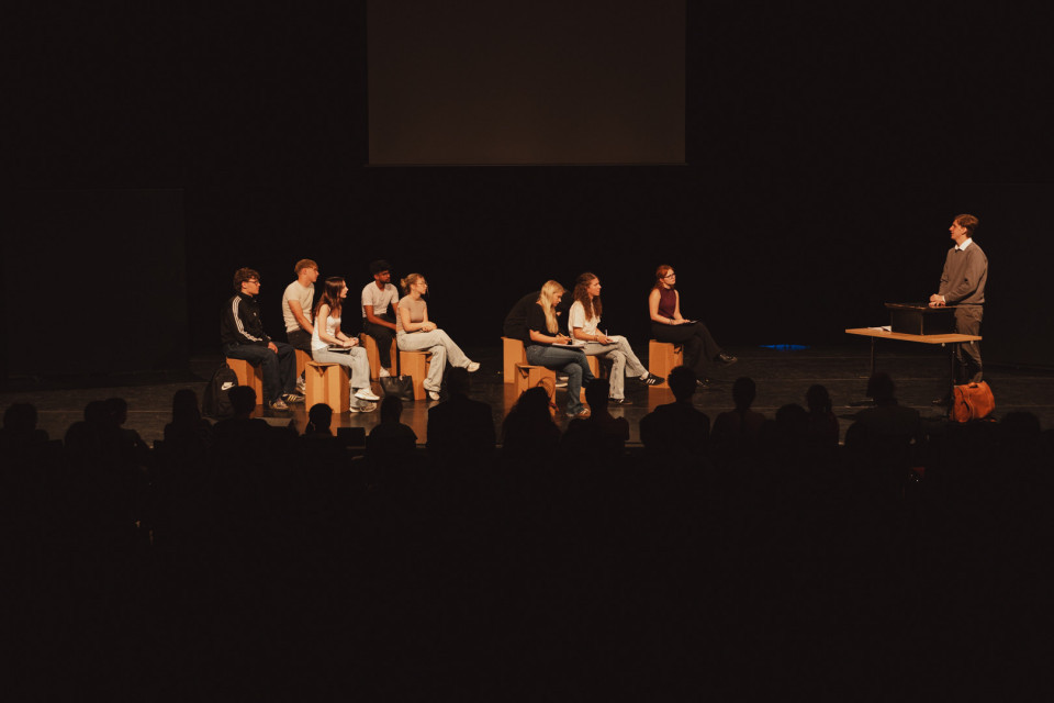 Auf einer dunklen Bühne sitzen mehrere junge Menschen auf Holzkisten und blicken zu einem stehenden Mann an einem Tisch rechts. Im Vordergrund sind die Silhouetten des Publikums zu sehen, während die Szene wie eine Theater- oder Präsentationssituation wirkt.