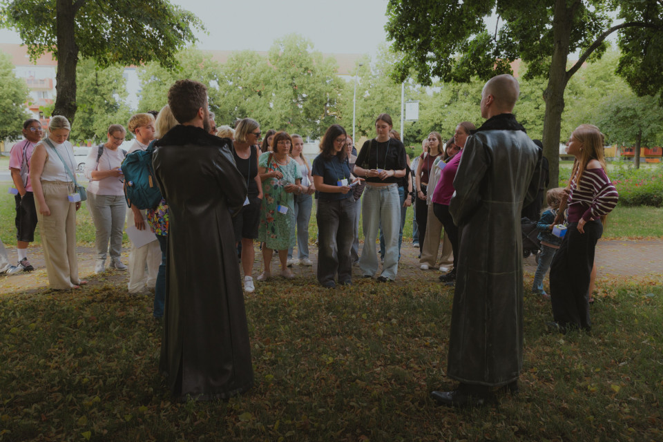 Eine Gruppe von Menschen steht in einem Park unter Bäumen und hört zwei Personen in langen schwarzen Mänteln zu. Die Teilnehmenden wirken wie bei einer Führung oder Veranstaltung und tragen teils Namensschilder sowie Notizen in den Händen.