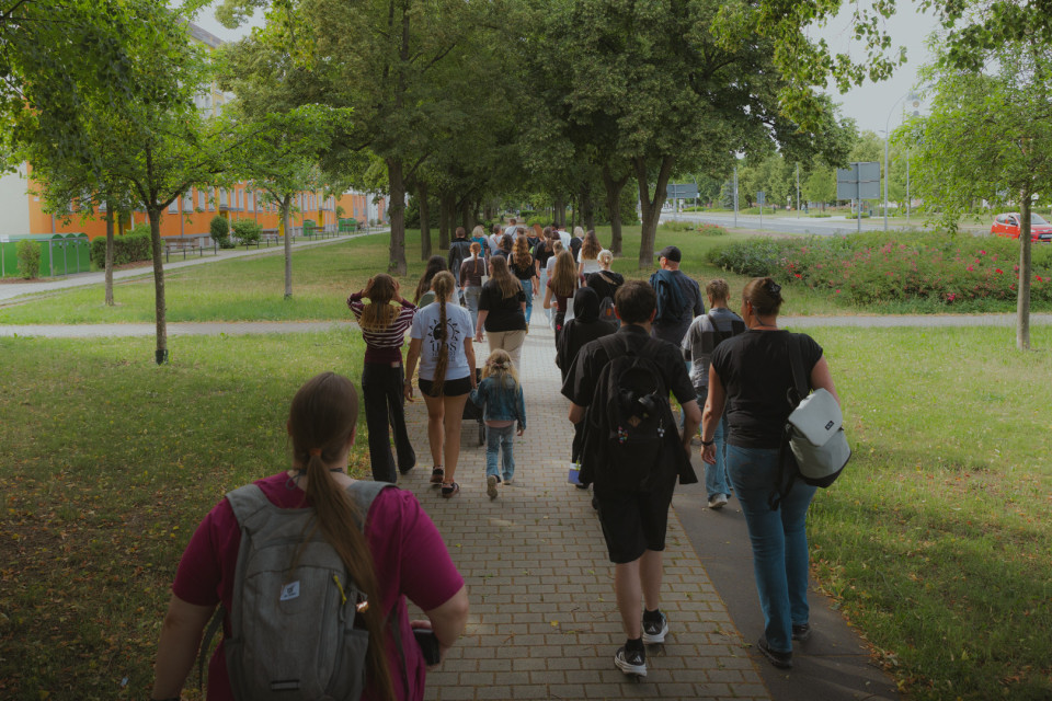 Eine Gruppe von Menschen geht auf einem gepflasterten Weg durch eine grüne, von Bäumen gesäumte Anlage. Links sind Wohngebäude zu sehen, rechts verlaufen eine Straße und blühende Büsche.