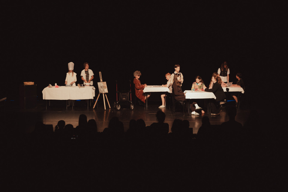 Auf einer dunklen Theaterbühne führen mehrere Kinder eine Szene mit gedeckten Tischen, Stühlen und Requisiten auf. Links steht ein Kind mit Kochmütze an einem Tisch, während rechts mehrere Kinder wie in einem Café oder Restaurant zusammensitzen.