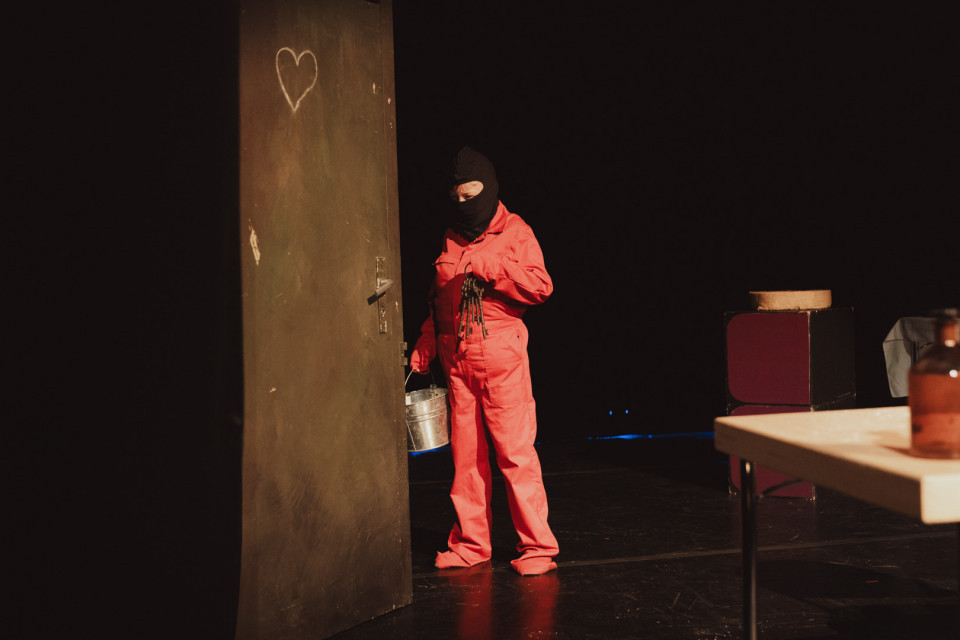 Eine Person in rotem Overall und schwarzer Gesichtsmaske steht neben einer großen Tür, auf der ein Herz gezeichnet ist. Die Szene wirkt wie eine dunkle Bühnenkulisse mit wenigen Requisiten und warmem Licht.