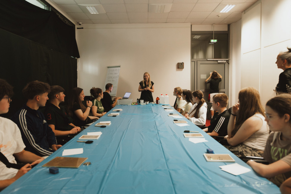 Eine Gruppe von Jugendlichen sitzt in einem Raum an einem langen, mit blauer Folie bedeckten Tisch und hört einer Frau zu, die vorne etwas erklärt. Auf dem Tisch liegen Materialien und kleine Gegenstände, während im Hintergrund weitere Personen bei der Vorbereitung oder Betreuung helfen.