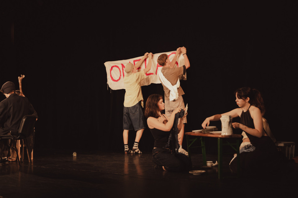 Auf einer dunklen Theaterbühne halten zwei Personen ein großes weißes Banner mit roten Buchstaben hoch. Rechts sitzen und knien weitere Darstellerinnen an einem kleinen Tisch mit Geschirr, während links eine Person mit dem Rücken zum Publikum auf einem Stuhl sitzt.
