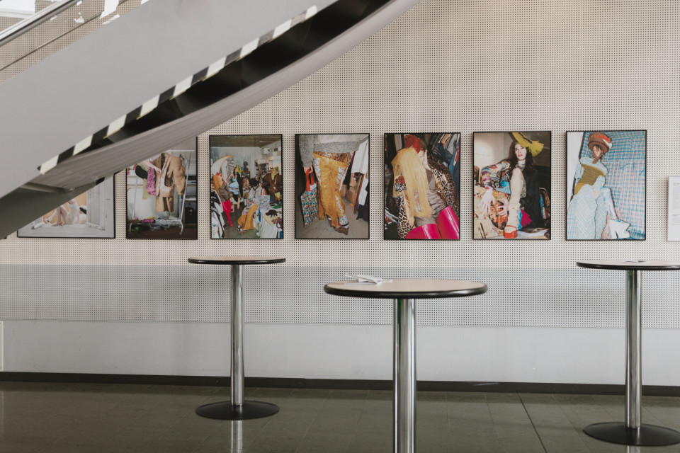 An einer perforierten Wand hängen mehrere gerahmte Modefotografien mit auffälligen Outfits und bunten Szenen. Im Vordergrund stehen runde Stehtische, während eine große Treppe diagonal durch den oberen Bildbereich verläuft.