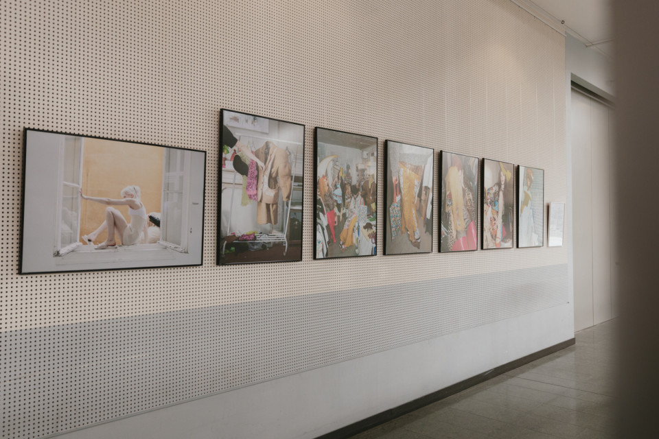 An einer perforierten weißen Galeriewand hängen mehrere gerahmte Fotografien in einer Reihe. Die Bilder zeigen überwiegend Mode- und Porträtszenen, während rechts ein heller Flur mit Tür zu sehen ist.