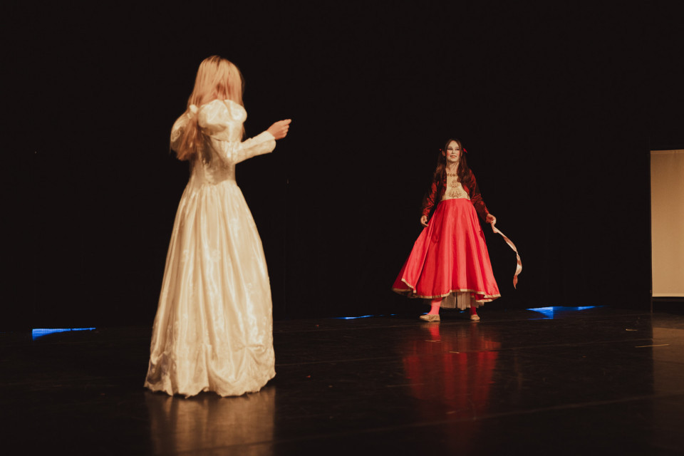 Auf einer dunklen Theaterbühne stehen zwei Mädchen in langen, festlichen Kleidern einander gegenüber. Im Vordergrund ist ein Mädchen in Weiß von hinten zu sehen, während das Mädchen in Rot weiter hinten ein Band in der Hand hält.