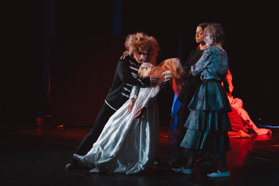 Auf einer dunkel beleuchteten Theaterbühne stützen mehrere kostümierte Darsteller eine Frau in einem weißen Kleid, die bewusstlos oder erschöpft nach vorne sinkt. Dramatisches rotes und blaues Licht sowie die auffälligen Masken und historischen Kostüme erzeugen eine unheimliche, spannungsvolle Szene.