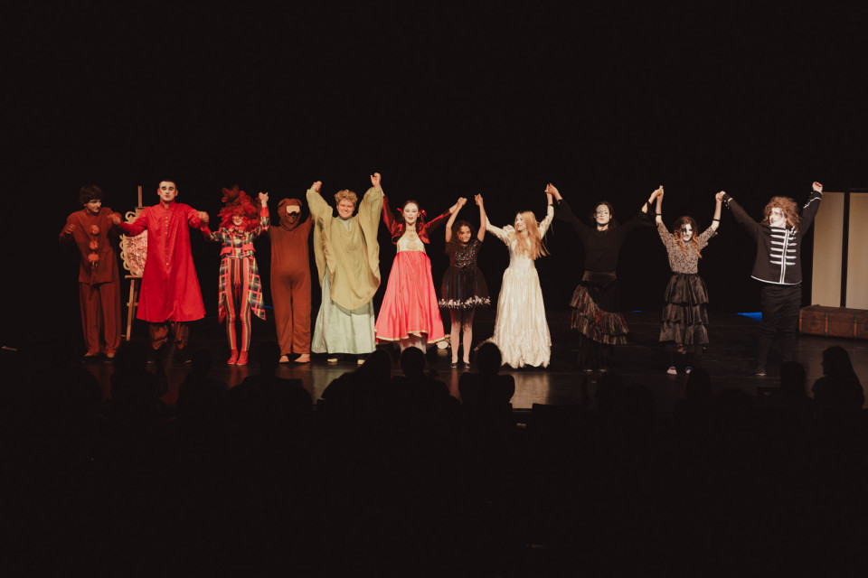 Eine Gruppe kostümierter Schauspieler steht auf einer dunklen Theaterbühne und hält sich nach einer Aufführung an den Händen. Vorne sind die Silhouetten des Publikums zu sehen, während die Darsteller gemeinsam Applaus entgegennehmen.