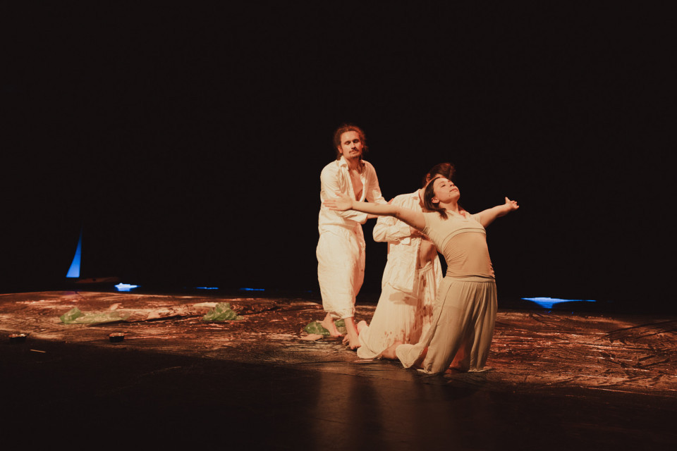 Auf einer dunklen Bühne führen drei Tänzer in hellen, beigen Kostümen eine ausdrucksstarke Choreografie auf. Warmes Licht hebt ihre Körperhaltungen hervor, während der Hintergrund fast vollständig schwarz bleibt.