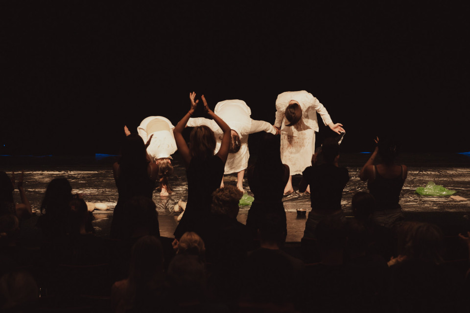Auf einer dunklen Theaterbühne verbeugen sich mehrere Darsteller in weißen Kostümen vor dem Publikum. Im Vordergrund stehen die Zuschauer als Silhouetten und applaudieren den Künstlern.