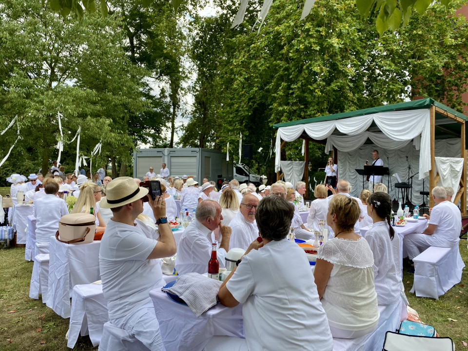 Viele Menschen in weißer Kleidung sitzen bei einer festlichen Veranstaltung im Freien an gedeckten Tischen unter Bäumen. Auf einer kleinen, mit weißen Stoffbahnen dekorierten Bühne treten Musiker vor dem Publikum auf.