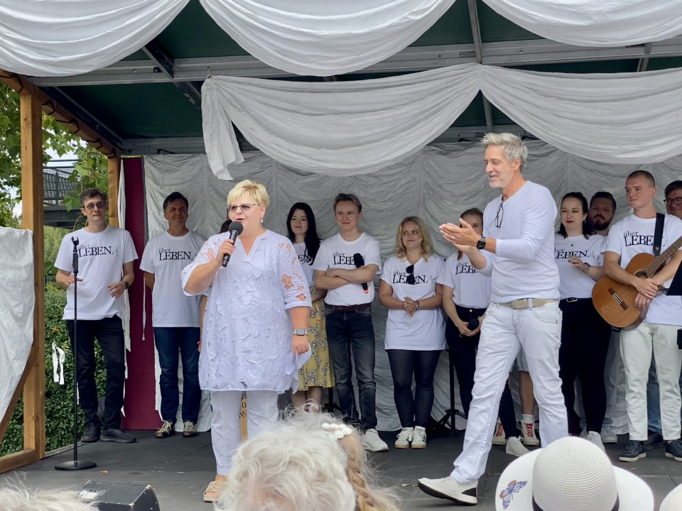 Auf einer kleinen Bühne spricht eine Frau in weißer Kleidung mit einem Mikrofon vor einem Publikum. Hinter ihr stehen mehrere Personen in weißen „über LEBEN“-T-Shirts, während ein Mann applaudiert und rechts ein Gitarrist zu sehen ist.