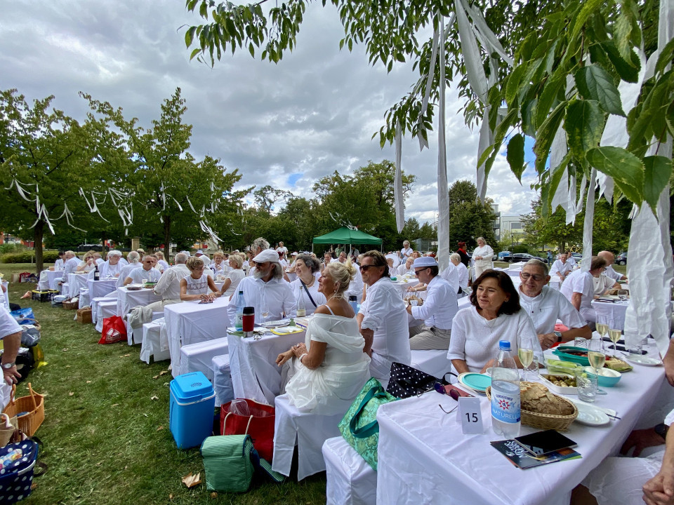 Viele Menschen in weißer Kleidung sitzen an langen, weiß gedeckten Tischen bei einem festlichen Picknick im Park. Zwischen den Bäumen hängen weiße Bänder, während ein bewölkter Himmel über der geselligen Veranstaltung liegt.