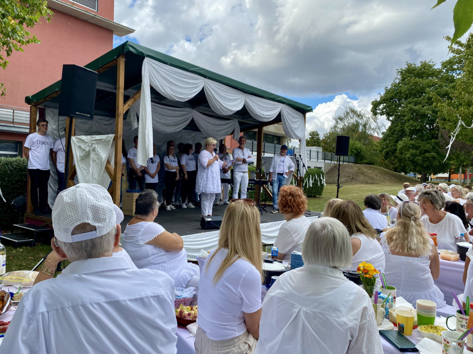 Eine Gruppe weiß gekleideter Menschen sitzt bei einer Veranstaltung im Freien vor einer kleinen geschmückten Bühne. Auf der Bühne spricht eine Frau ins Mikrofon, während weitere Personen und Musikausrüstung im Hintergrund zu sehen sind.