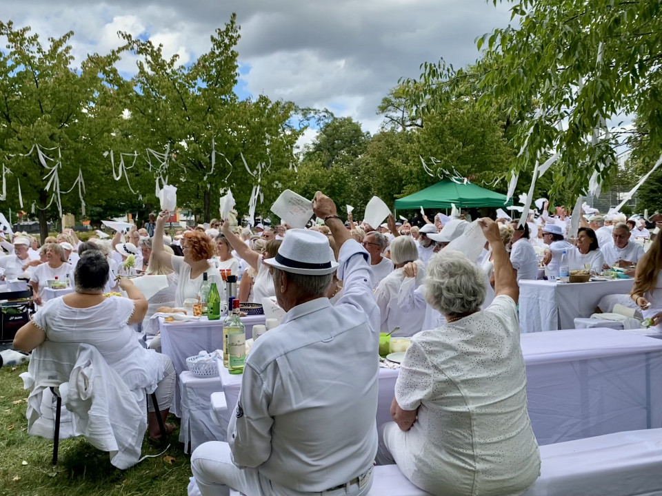 Viele Menschen in weißer Kleidung sitzen an langen, weiß gedeckten Tischen in einem Park und heben Servietten oder Tücher in die Luft. Zwischen den grünen Bäumen hängen weiße Bänder, während ein bewölkter Himmel über der festlichen Zusammenkunft liegt.