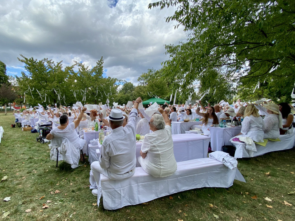 Eine große Gruppe von Menschen in weißer Kleidung sitzt an weiß gedeckten Tischen in einem Park und schwenkt Servietten oder Tücher. Über ihnen hängen Wolken am Himmel, während grüne Bäume den festlichen Außenbereich umrahmen.
