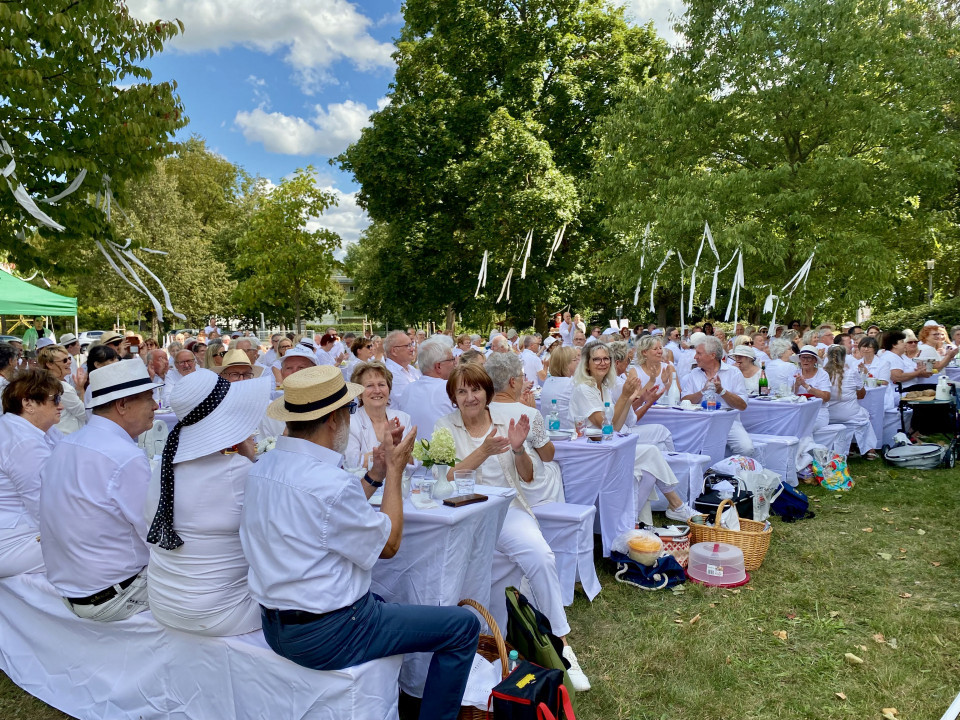 Eine große Gruppe festlich in Weiß gekleideter Menschen sitzt an gedeckten Tischen in einem grünen Park und applaudiert. Über ihnen hängen weiße Bänder in den Bäumen, während bei sonnigem Wetter eine gesellige Feier im Freien stattfindet.