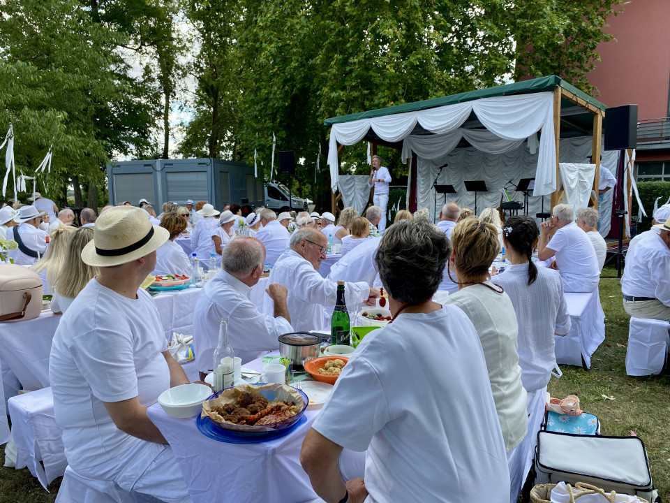 Viele weiß gekleidete Menschen sitzen im Freien an langen Tischen und hören einer Person auf einer geschmückten Bühne zu. Die Veranstaltung findet unter Bäumen statt, mit weißen Stoffdekorationen, Lautsprechern und gedeckten Tischen voller Speisen und Getränke.