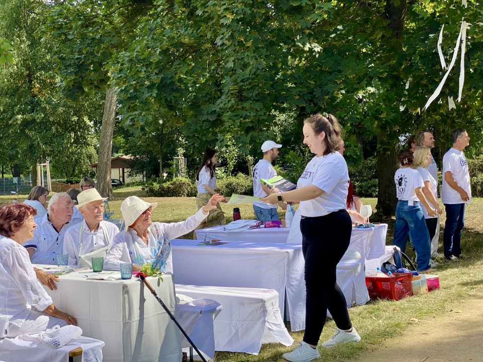 In einem grünen Park sitzen mehrere ältere Menschen in weißer Kleidung an festlich gedeckten Tischen, während eine junge Frau ihnen Flyer oder Unterlagen reicht. Im Hintergrund stehen weitere Personen in weißen T-Shirts zwischen den Bäumen, und die Szene wirkt wie eine sommerliche Gemeinschaftsveranstaltung im Freien.