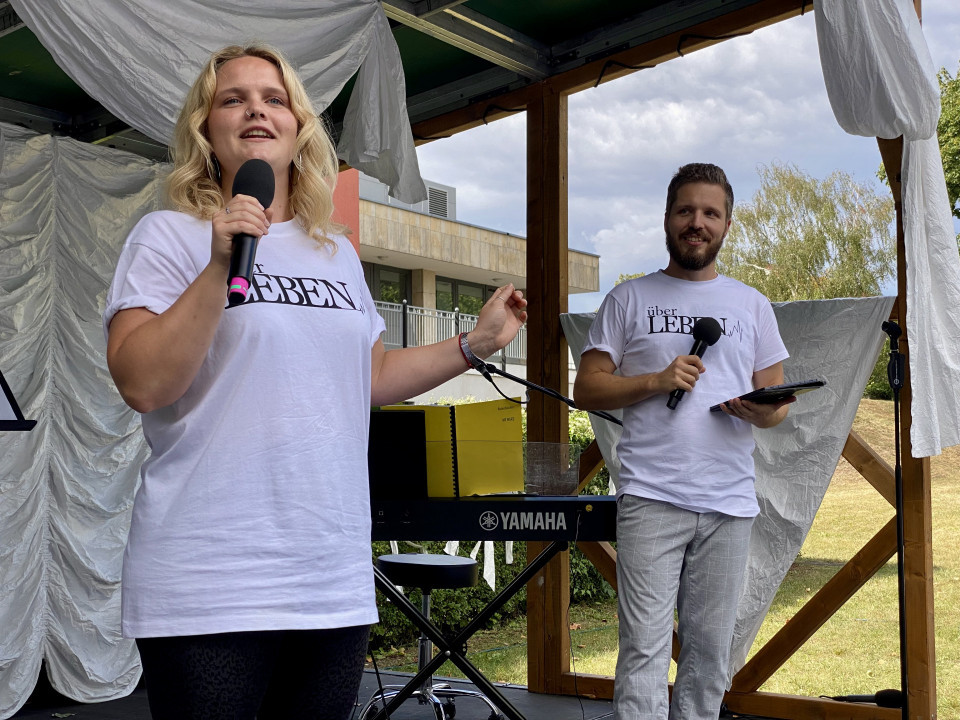 Eine blonde Frau steht auf einer kleinen Außenbühne und spricht mit einem Mikrofon, während sie ein weißes T-Shirt trägt. Neben ihr hält ein Mann ebenfalls ein Mikrofon und ein Klemmbrett; im Hintergrund sind weiße Stoffbahnen, ein Yamaha-Keyboard und ein bewölkter Himmel zu sehen.