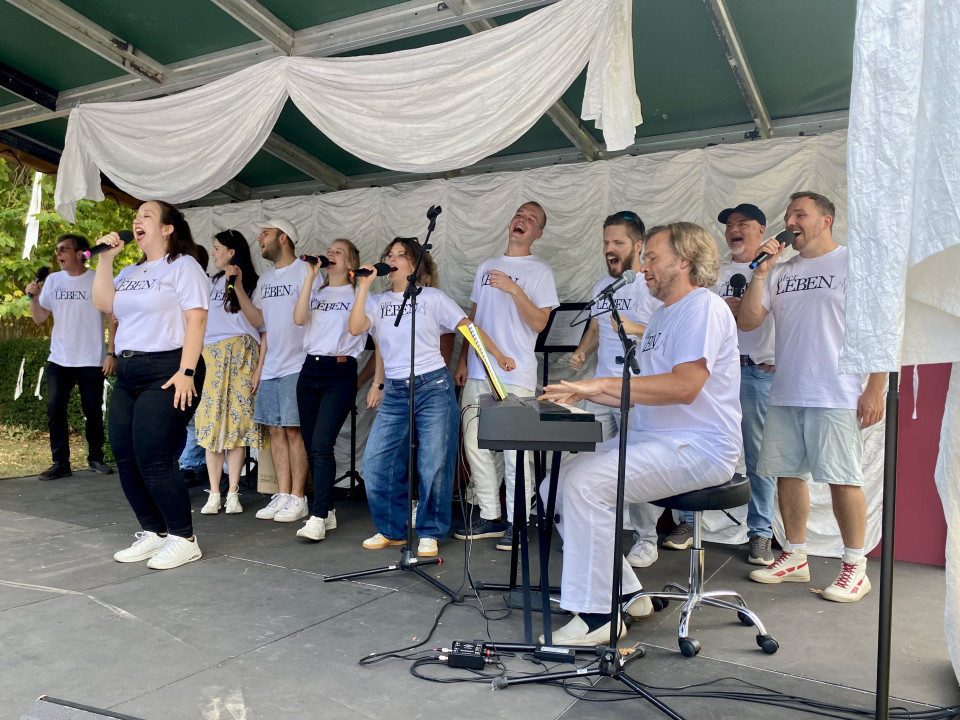 Eine Gruppe von Sängerinnen und Sängern steht auf einer überdachten Bühne und singt mit Mikrofonen, während ein Mann am Keyboard spielt. Alle tragen weiße T-Shirts, und die Bühne ist mit weißen Stoffbahnen dekoriert.