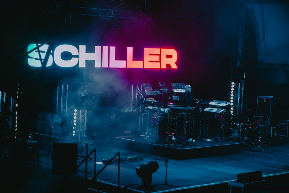 Auf einer dunklen Konzertbühne leuchtet im Hintergrund der große Schriftzug „SCHILLER“ in Blau, Weiß und Rot. Rechts sitzt ein Musiker zwischen Synthesizern, Keyboards und Schlagzeug, umgeben von Nebel und atmosphärischem Bühnenlicht.