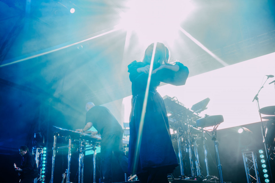 Auf einer Konzertbühne steht eine Sängerin im Gegenlicht, während grelle Scheinwerfer und blauer Nebel die Szene dominieren. Im Hintergrund spielen weitere Musiker an Gitarren, Schlagzeug und elektronischen Instrumenten.