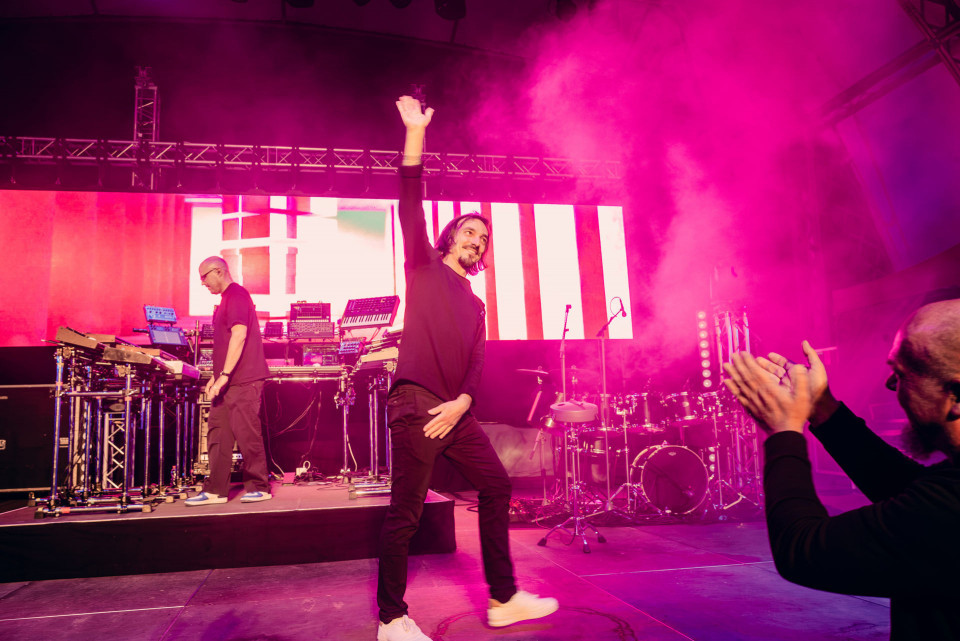 Auf einer Konzertbühne winkt ein Mann in schwarzer Kleidung ins Publikum, während pinker Rauch und helle LED-Wände die Szene beleuchten. Links steht ein Musiker an elektronischen Instrumenten, rechts klatscht ein Zuschauer vor einem Schlagzeug im Hintergrund.