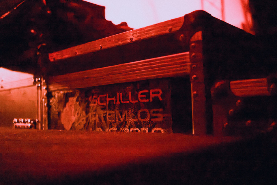 Das Foto zeigt eine dunkle, abgenutzte Transportkiste oder Bühnenausrüstung in intensiv rotem Licht. Auf der Vorderseite sind teilweise lesbare Schriftzüge und metallische Beschläge zu erkennen, während der Hintergrund unscharf bleibt.