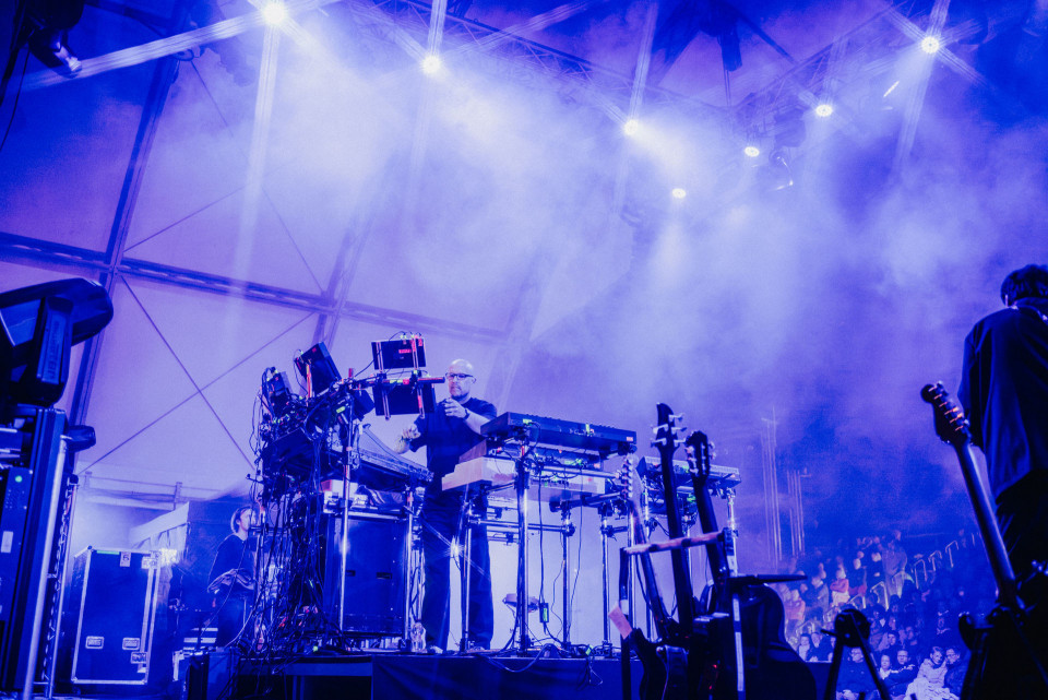 Auf einer Konzertbühne steht ein Musiker zwischen zahlreichen Synthesizern, Kabeln und technischen Geräten, umgeben von dichtem Nebel und blau-violettem Licht. Rechts sind Gitarren und ein weiterer Musiker zu sehen, während im Hintergrund das Publikum im Halbdunkel sitzt.