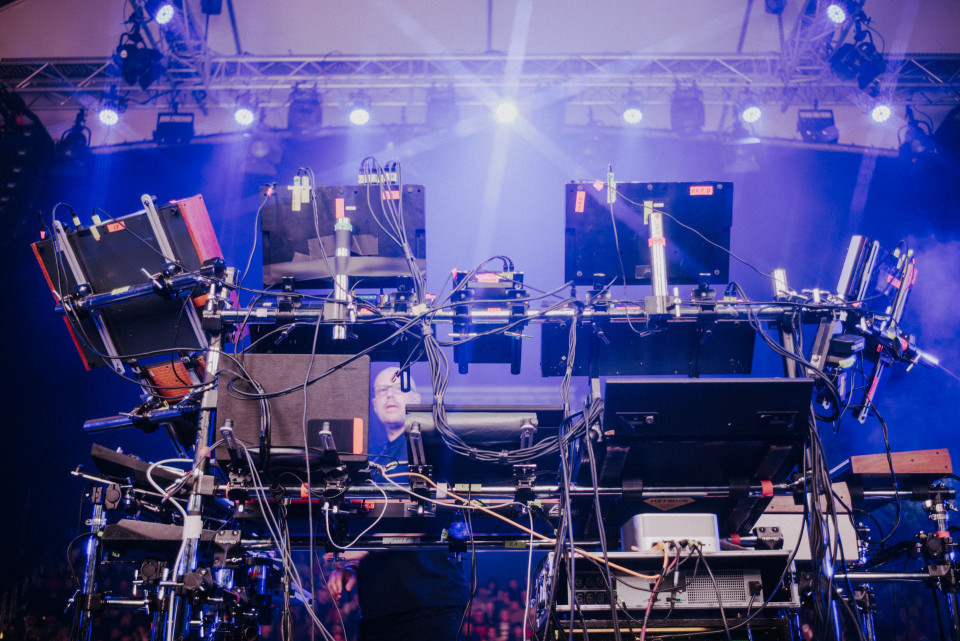 Auf einer Bühne steht ein komplexes elektronisches Musik-Setup mit vielen Monitoren, Kabeln, Halterungen und Steuergeräten. Im Hintergrund leuchten blaue und violette Scheinwerfer durch Nebel, während ein Musiker teilweise hinter der Technik zu sehen ist.
