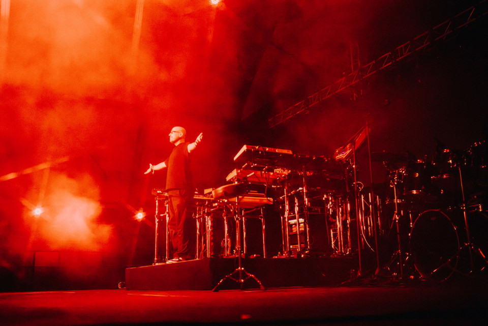 Ein Musiker steht mit ausgebreiteten Armen auf einer Bühne, die in intensives rotes Licht und dichten Nebel getaucht ist. Um ihn herum sind Keyboards, Percussion und ein Schlagzeug aufgebaut, während Scheinwerfer die dramatische Konzertatmosphäre verstärken.