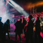 Eine große Menschenmenge sitzt und steht bei einem nächtlichen Konzert vor einer hell beleuchteten Bühne. Bunte Scheinwerfer, Nebel und kräftige Lichtstrahlen tauchen die Szene in eine lebendige Atmosphäre.