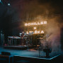 Auf einer dunklen Konzertbühne steht ein Musiker zwischen Synthesizern und elektronischem Equipment, während Nebel und blau-oranges Licht die Szene füllen. Im Hintergrund leuchtet groß der Schriftzug „SCHILLER x LIZÉKA“, daneben sind Schlagzeug, Mikrofone und weitere Bühneninstrumente zu sehen.