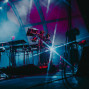 Auf einer Konzertbühne stehen zahlreiche elektronische Instrumente, Synthesizer und Kabel im dichten Nebel. Blaues und pinkes Licht sowie helle Scheinwerferstrahlen erzeugen eine dramatische, futuristische Atmosphäre.