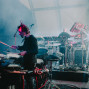 Ein Schlagzeuger spielt auf einer neblig beleuchteten Konzertbühne, umgeben von Mikrofonen, Kabeln und Percussion-Instrumenten. Im Hintergrund bedient ein weiterer Musiker elektronische Geräte und Synthesizer, während farbige Scheinwerfer durch den Bühnenrauch strahlen.